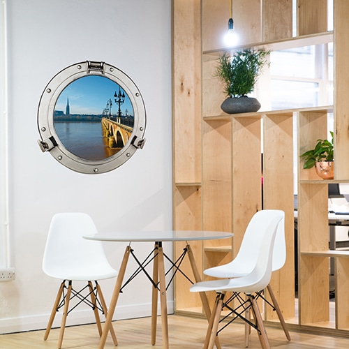 Salle à manger avec claustra en bois illuminée par un faux hublot de bateau adhésif offrant la vue sur le Pont de Pierre à Bordeaux.