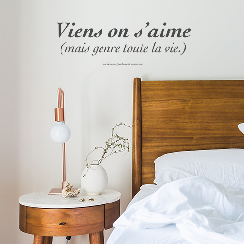 Sticker adhésif Vient on s'aime citation posée au dessus d'une tête de lit avec une lampe