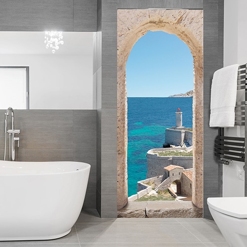 Dans une salle de bain moderne grise et blanche on s'évade à l'aide de cet adhésif pour porte qui nous emmène au bord de la Méditerranée dans une vieux village entouré d'une forteresse